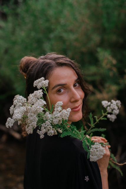 A woman with her hair in a bun looks over her shoulder while holding white yarrow flowers in a dark green outdoor setting.