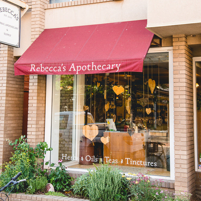 A storefront for Rebecca's Apothecary with a red awning, brick building exterior, large glass windows, and heart decorations hanging in the window display.
